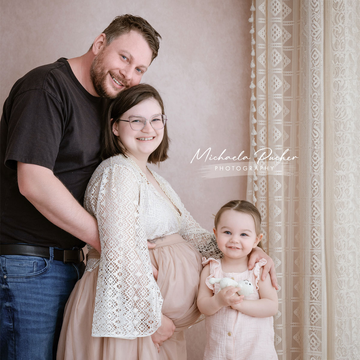 Familie mit werdender Mama, Papa und kleinem Kind posiert lächelnd im Studio, alle eng aneinandergeschmiegt.